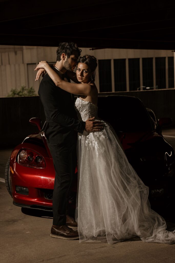 bride and groom embrace in front of red sportscar during urban wedding portraits.