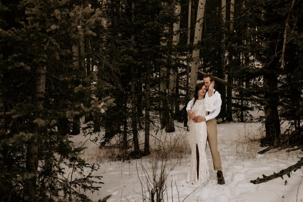 bride and groom stand in forest and snow during adventure elopement photo shoot.