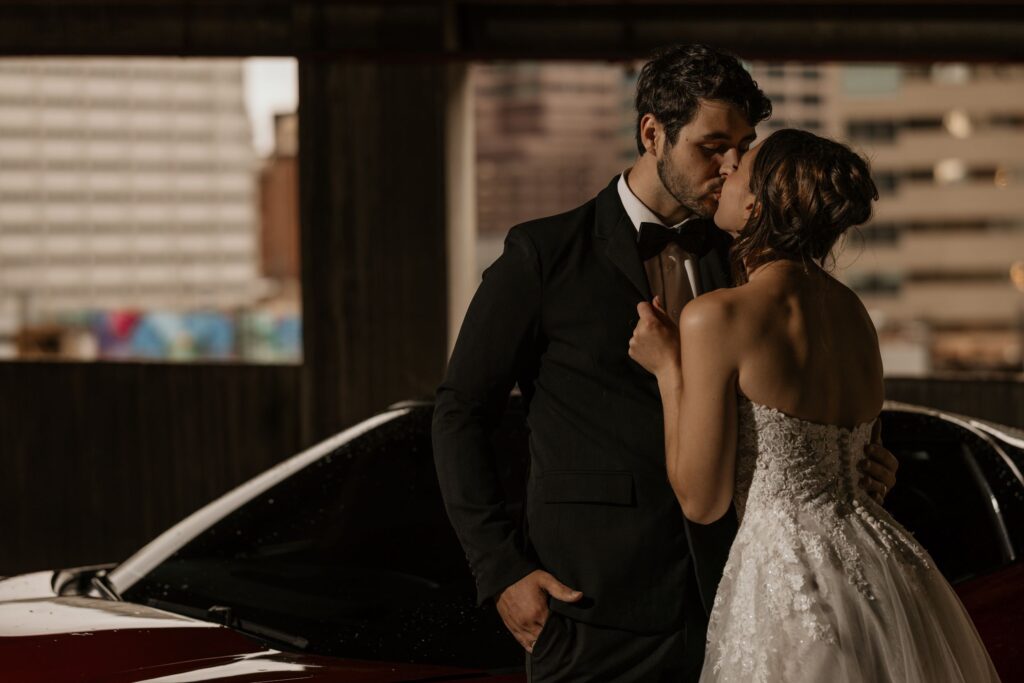 bride grabs groom and kisses him beside car during denver wedding portraits.