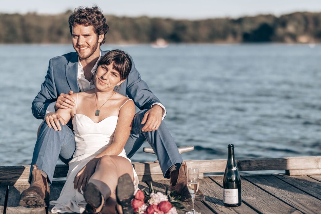 bride and groom sit with a bottle of champagne on dock during lake elopement in colorado.