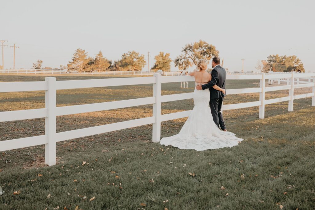 Bride and groom stand by white fence overlooking horse pasture, during rustic wedding portraits.