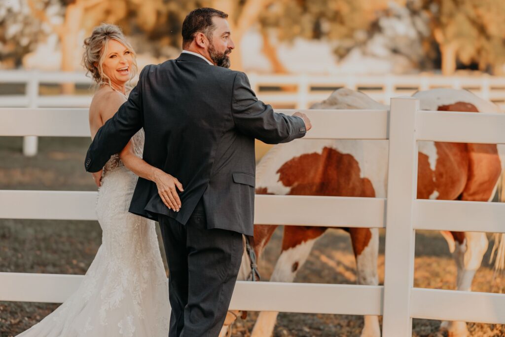 bride and groom smile, standing at white fence by a horse, during western wedding photos.