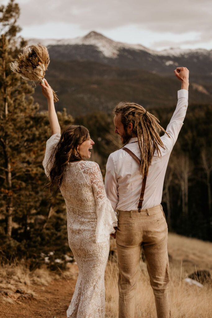 bride and groom throw their fists up in celebration, mountains in the background, during rustic mountain wedding.