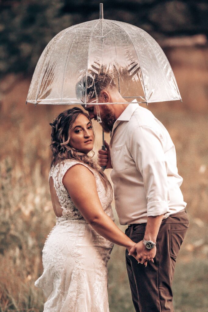 bride and groom stand under clear umbrella during farmhouse wedding portraits with colorado wedding photographer.
