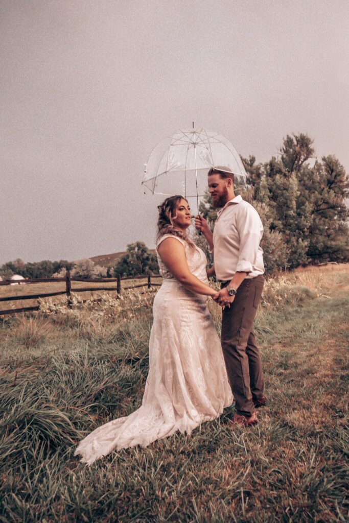 bride and groom stand under clear umbrella during rustic wedding in colorado.