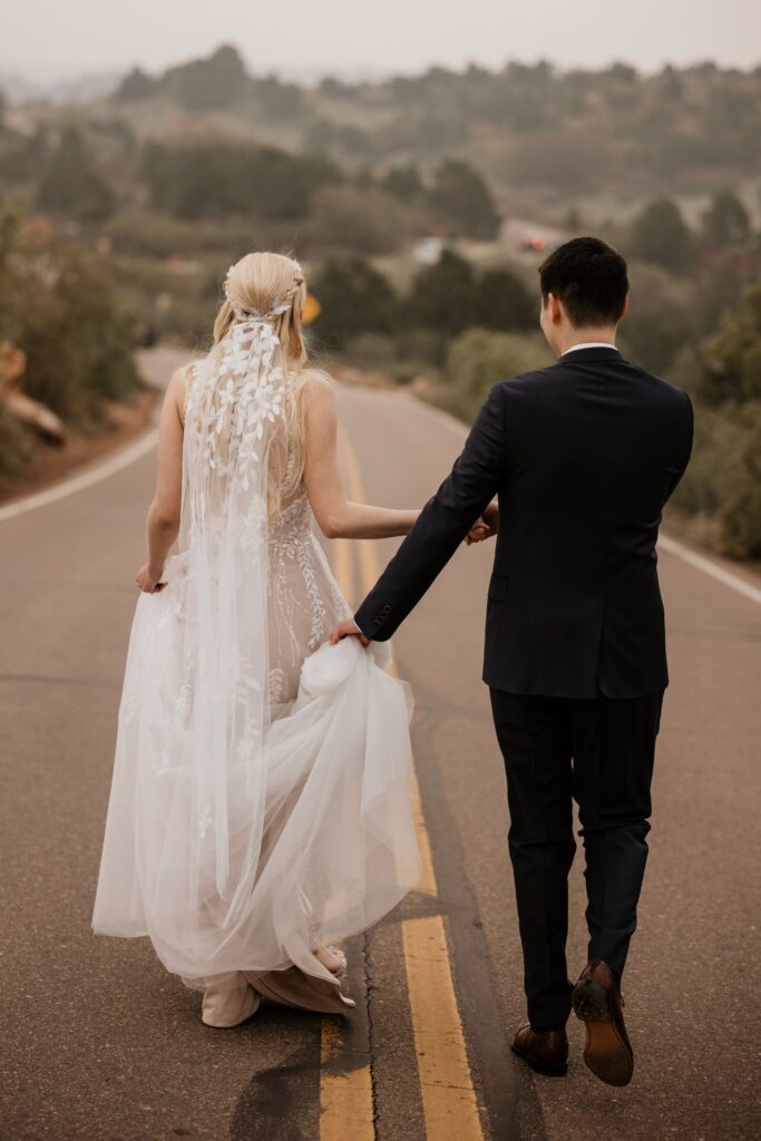 groom holds brides dress as they walk across the road at Colorado Springs' Garden of the Gods during wedding photos.