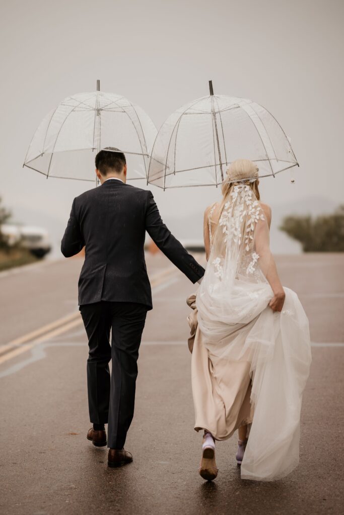 couple holds umbrellas as they walk across the road at Colorado Springs' Garden of the Gods during wedding photos.