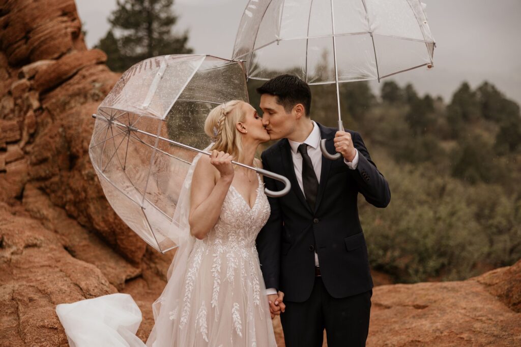 bride and groom kiss under clear umbrellas during a rainy wedding photo shoot at garden of the gods colorado.