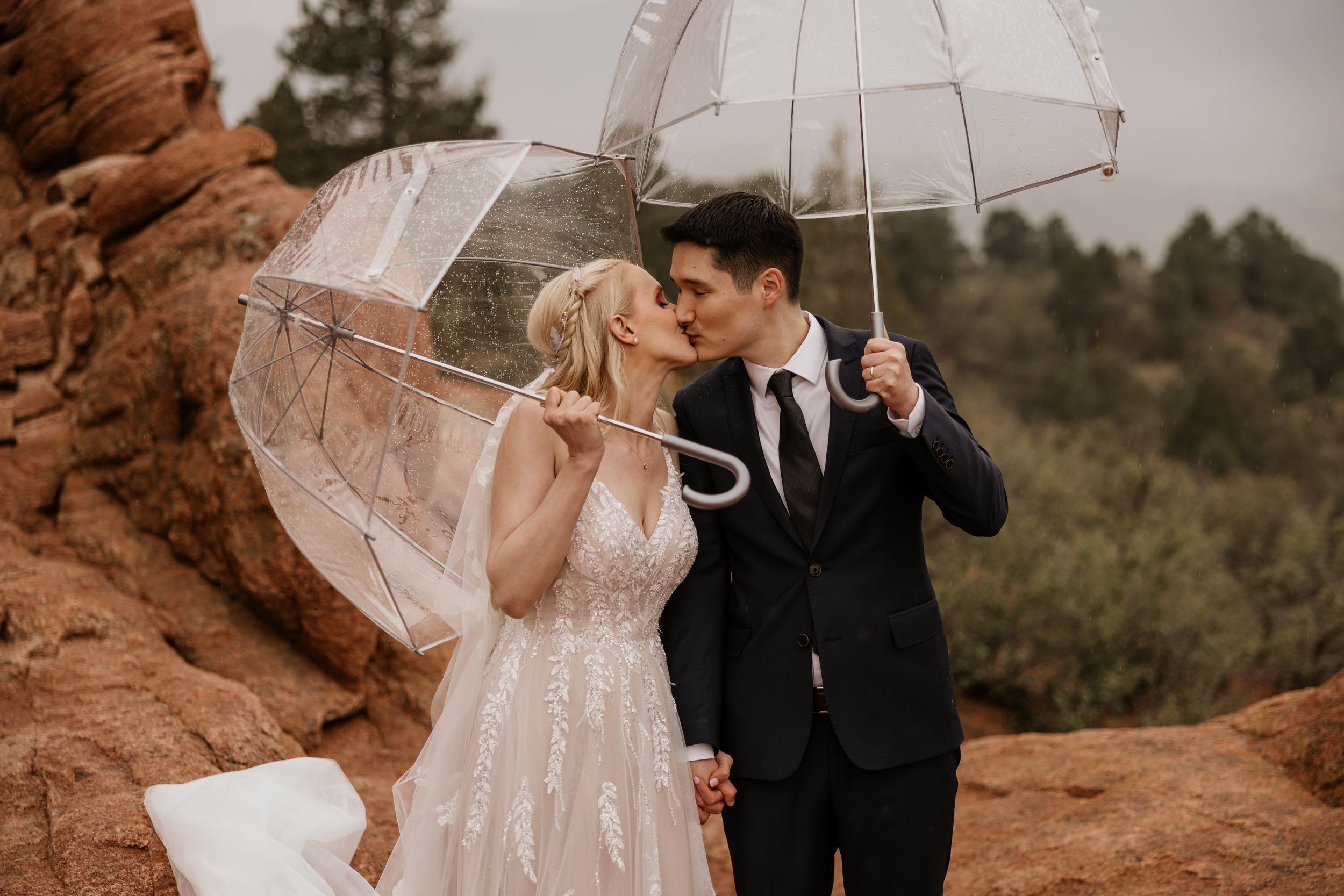 bride and groom kiss under clear umbrellas during a rainy wedding photo shoot at garden of the gods elopement colorado.