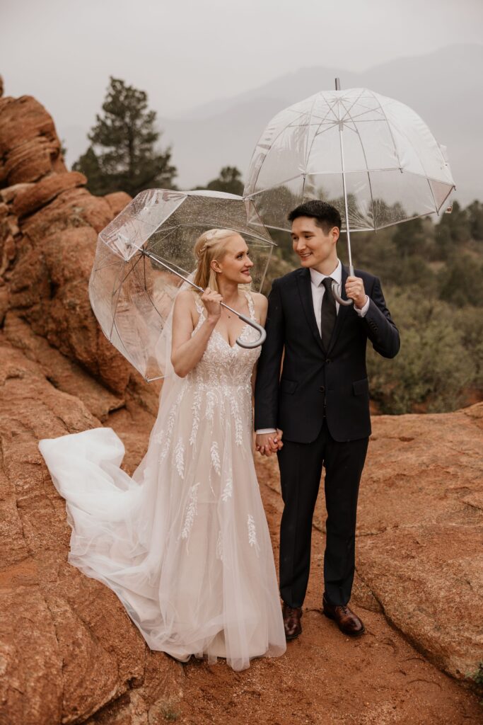 bride and groom hold clear umbrellas during rainy wedding photo session in garden of the gods colorado.
