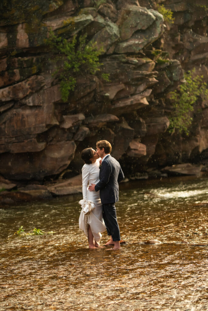 bride and groom kiss in the rain, standing in a stream during their micro wedding at river bend venue in lyons colorado.