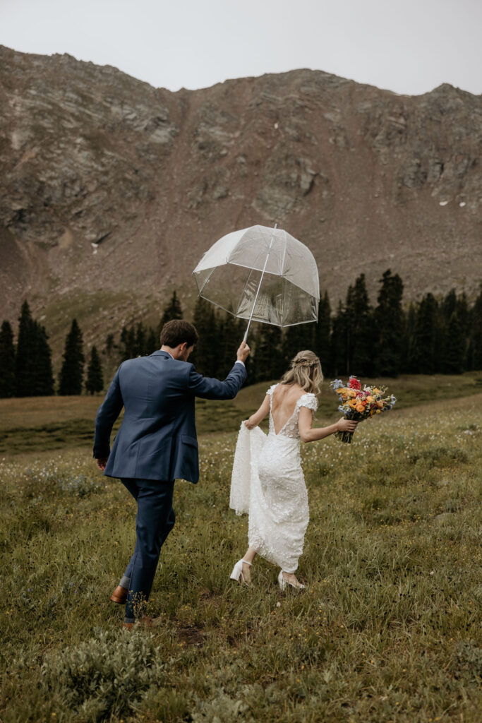 bride and groom walk out into rainy field with umbrella during their ski wedding in the colorado mountains.