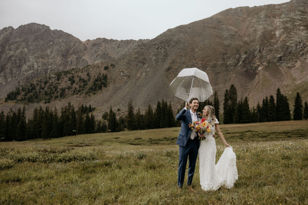 bride and groom stand outside under clear umbrella during their ski mountain wedding in colorado.