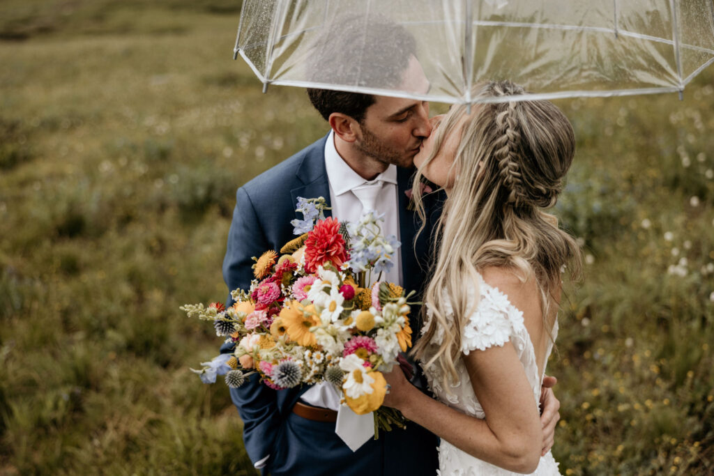 bride and groom kiss under clear umbrella during their ski mountain wedding in colorado.