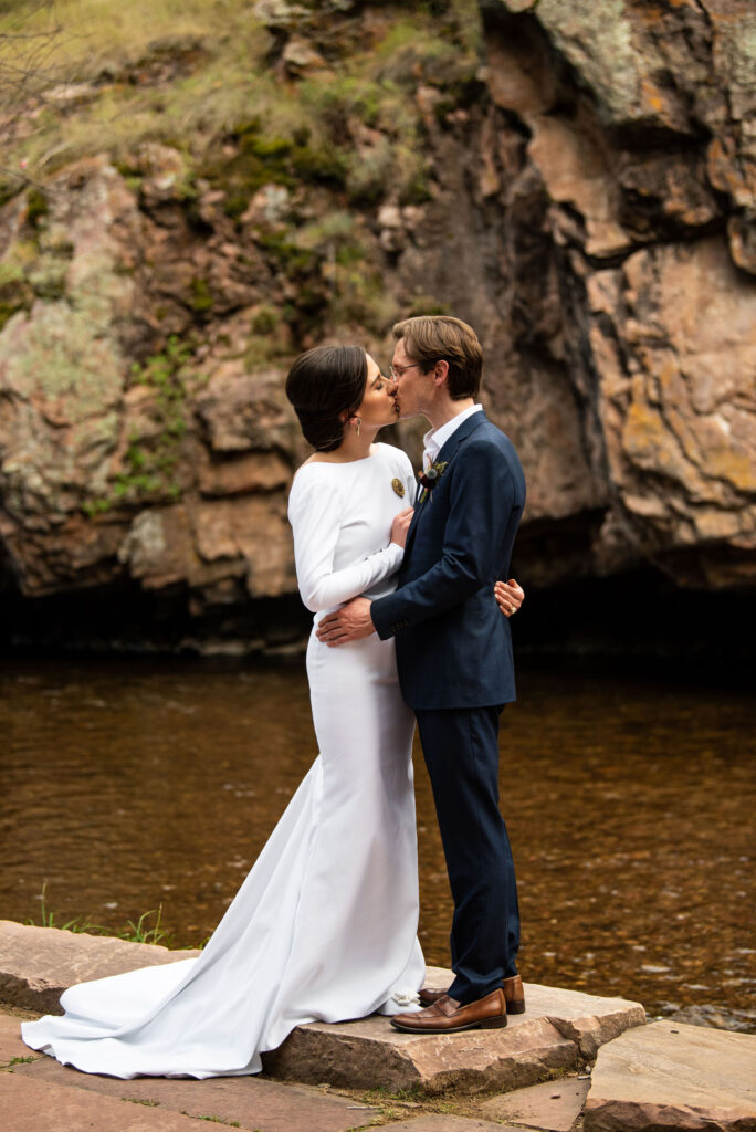 bride and groom kiss on the riv ers edge during their micro wedding in lyons colorado.