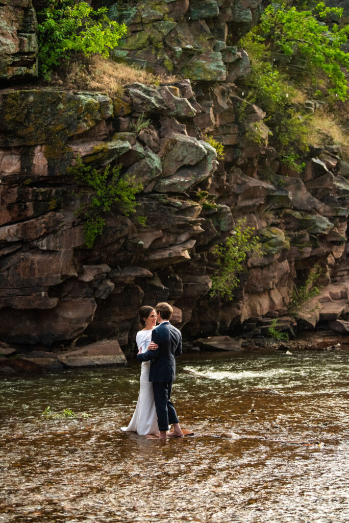 bride and groom stand in the rain, in a stream during their micro wedding at river bend venue in lyons colorado.