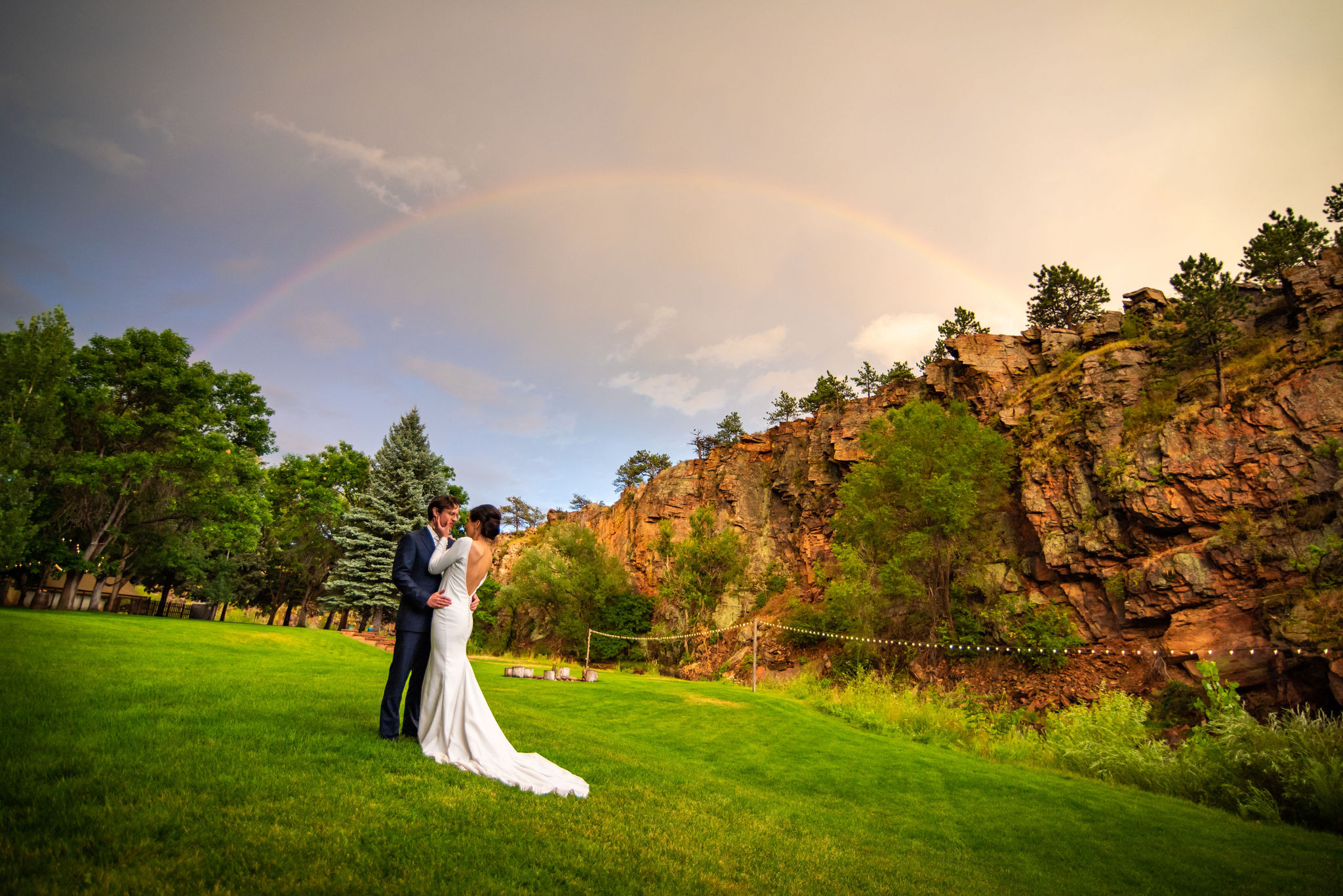 bride and groom stand and embrace under rainbow at river bend wedding venue.