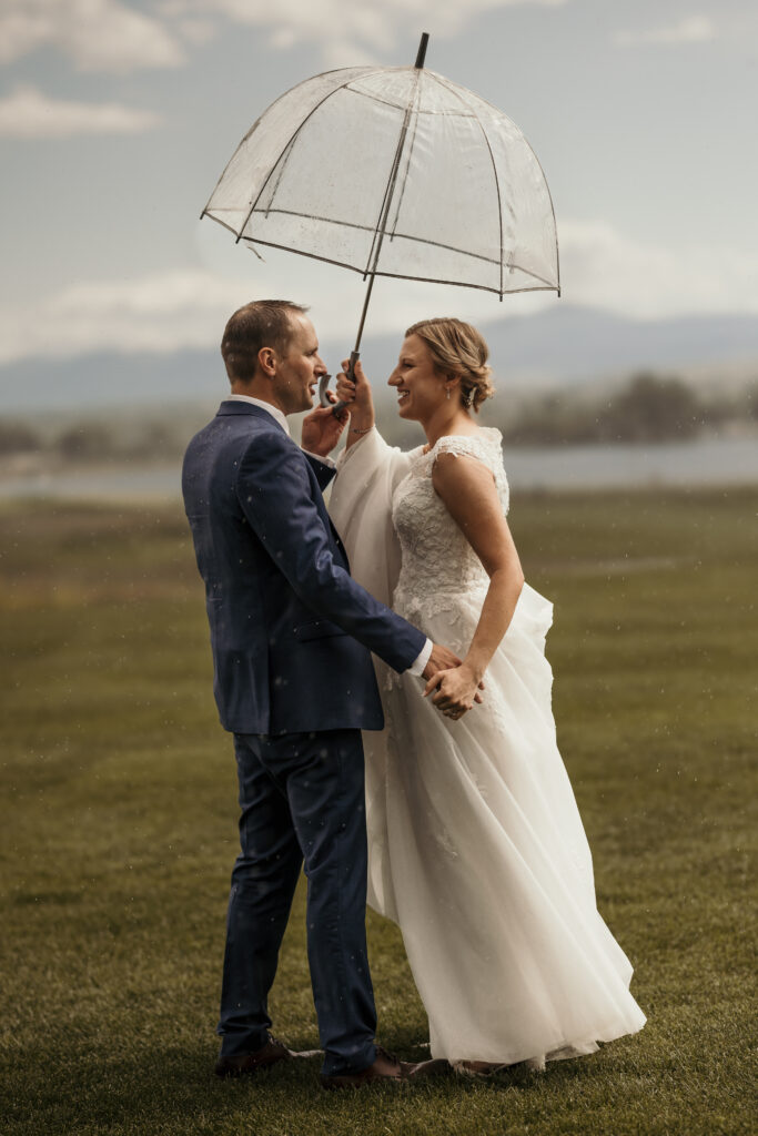 Bride and groom stand under clear umbrella in the rain, during spring micro wedding in colorado.