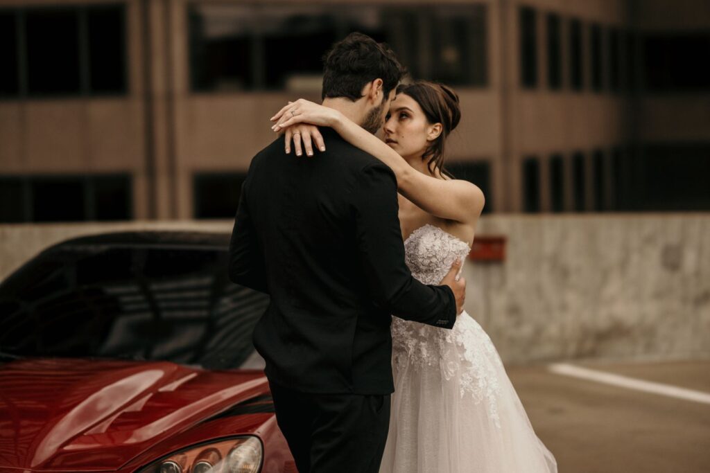 bride wraps arms around grooms neck beside red sportscar during urban bridal portrait photos.