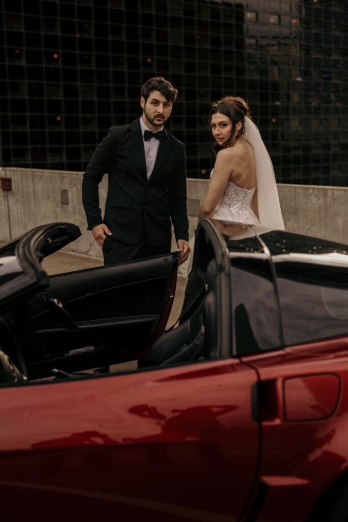 bride and groom stand beside front of red sportscar, during wedding portraits in denver colorado.