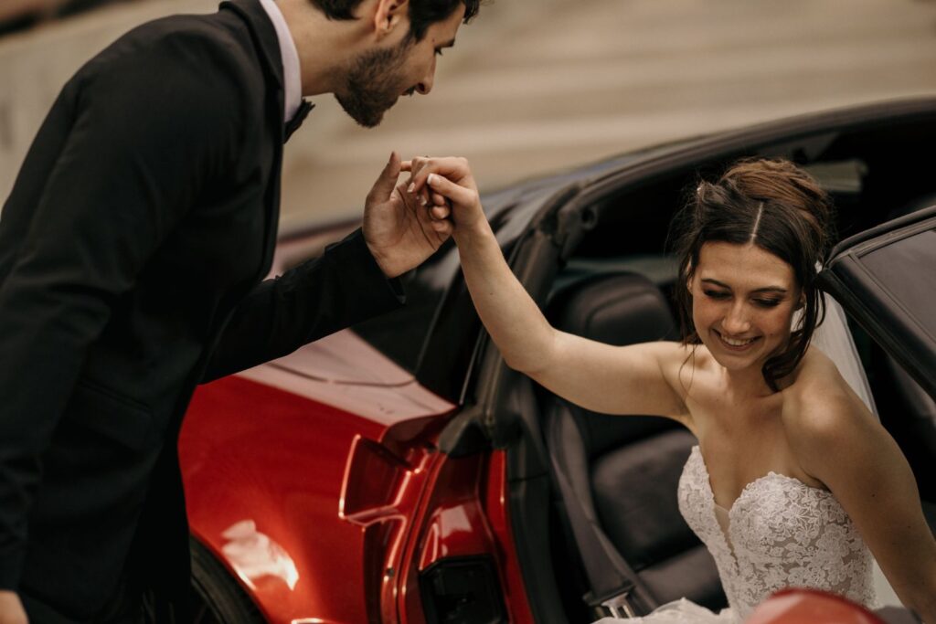 groom helps bride out of car and kisses her hand during urban bridal portrait photos.