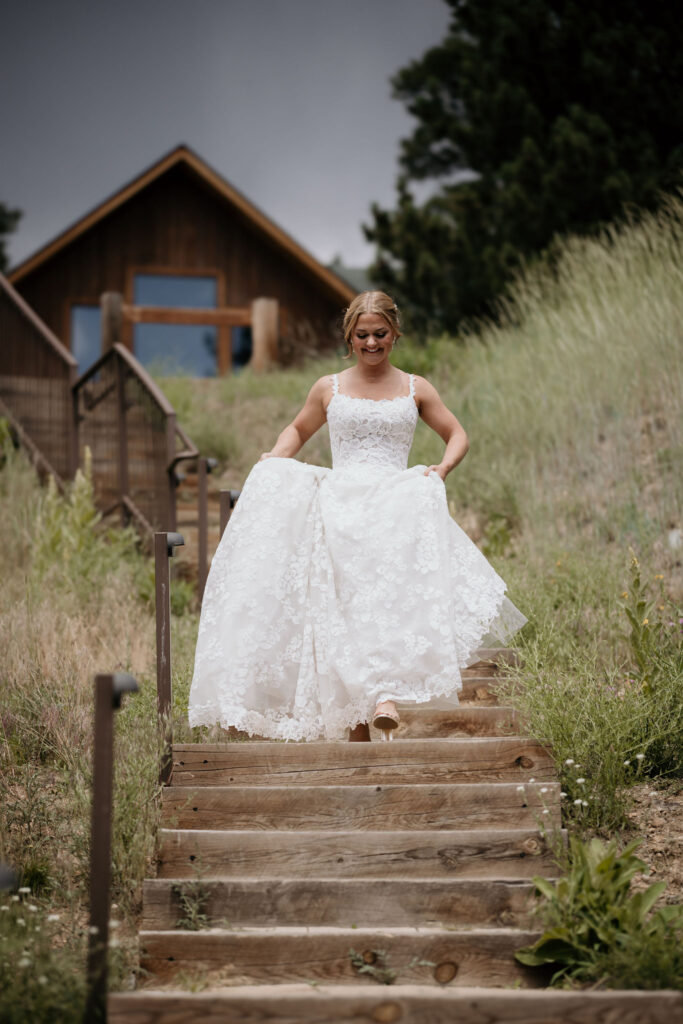 bride holds dress and walks down steps while preparing for first look at skyview wedding venue in estes park colorado.
