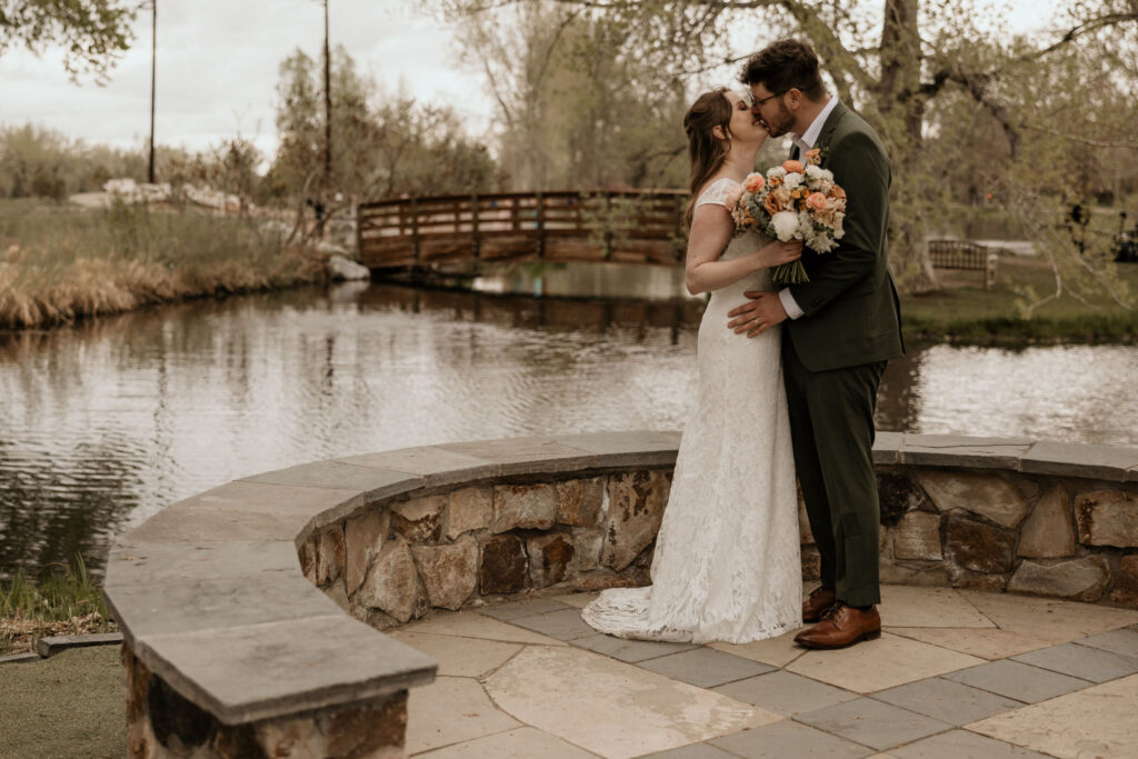 bride and groom kiss in front of pond with bridge, during spring micro wedding in colorado.