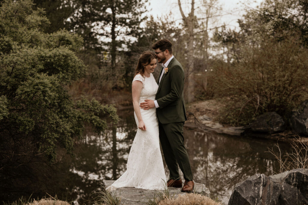 newly wed couple stand on rock in front of pond in the garden during spring colorado micro wedding.