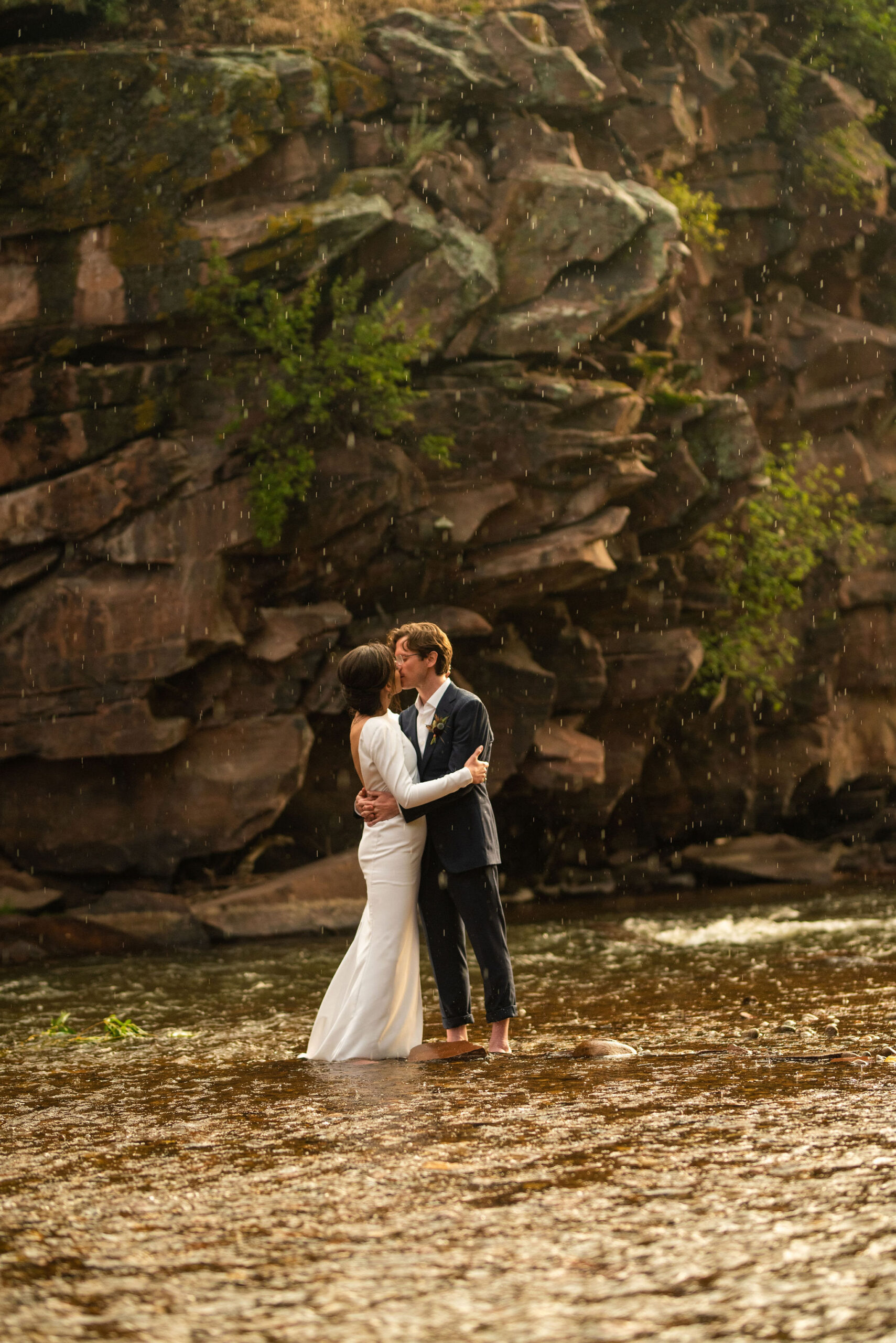 bride and groom kiss in the rain, standing in a stream during their micro wedding at river bend venue in colorado.
