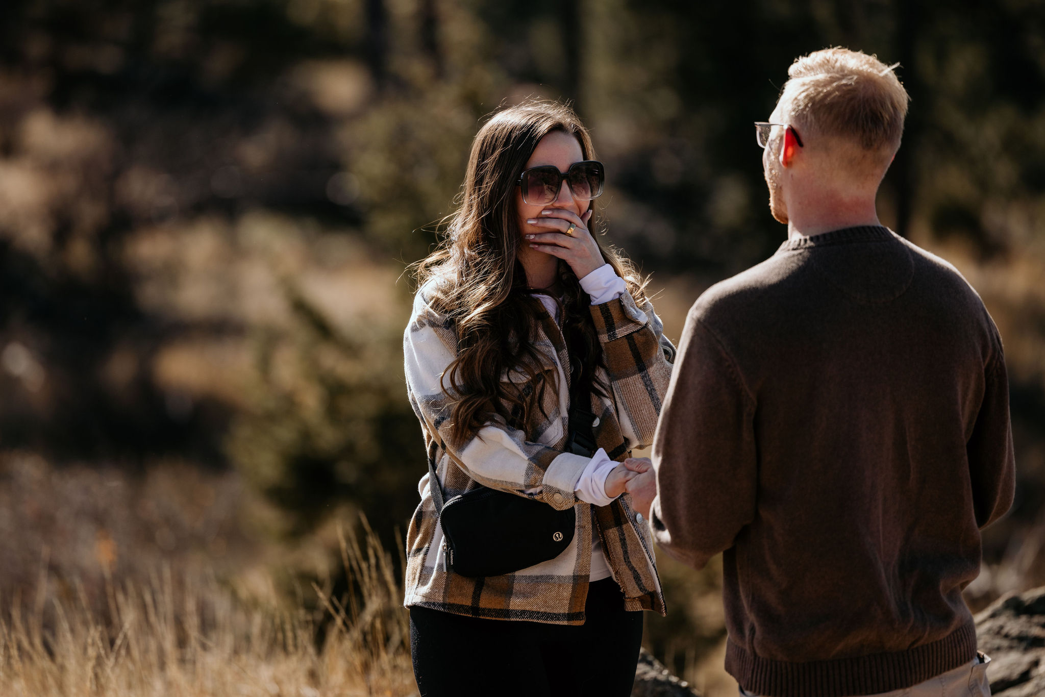 woman covers mouth as she realizes her boyfriend is about to propose while hiking in the mountains.