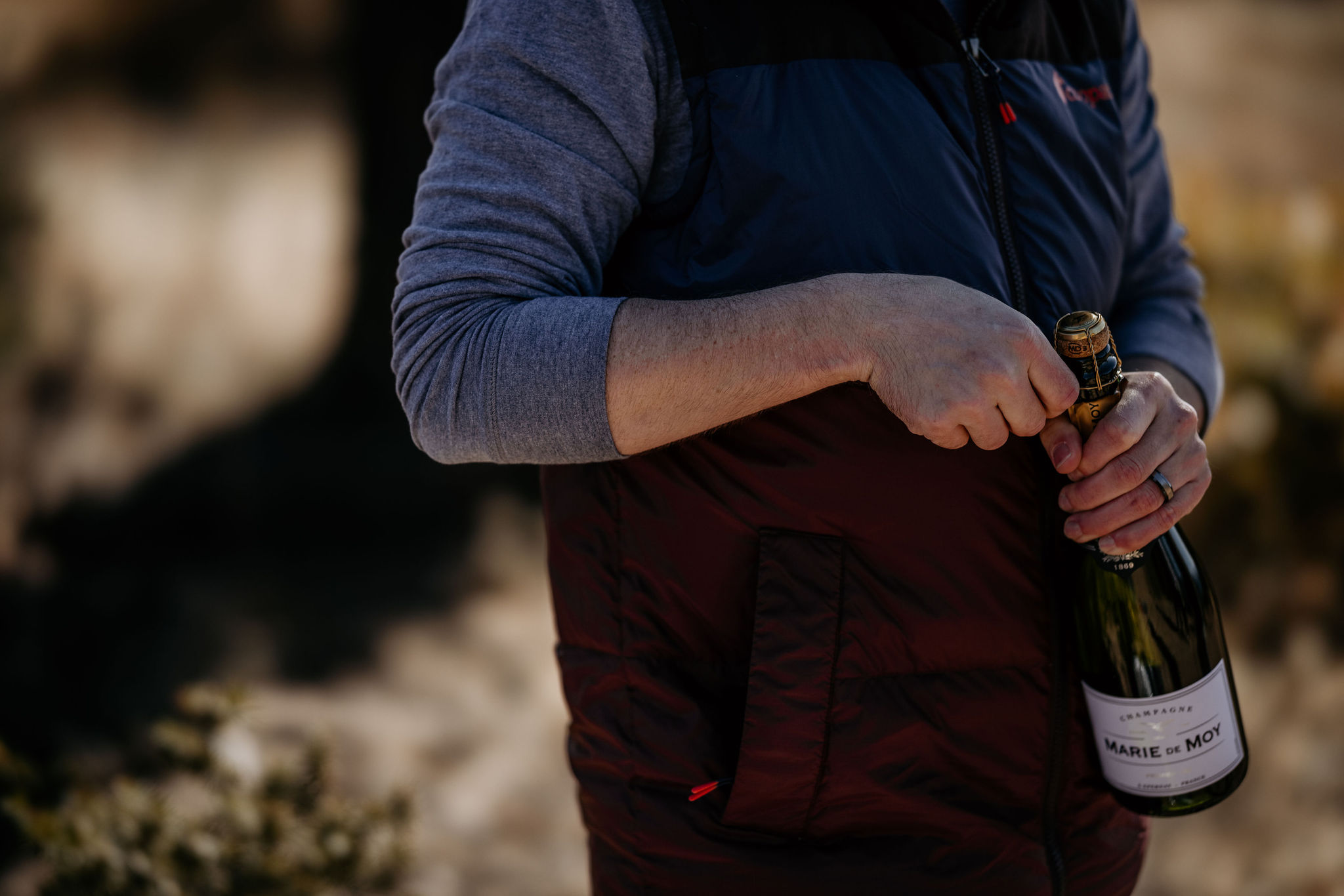 man opens bottle of champagne while celebrating a proposal in the mountains.