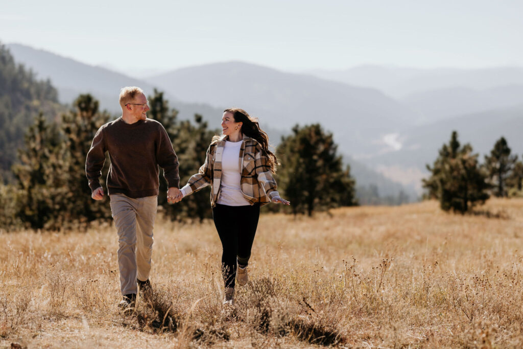 man and woman run hand in hand through a field in the colorado mountains for engagement photos.