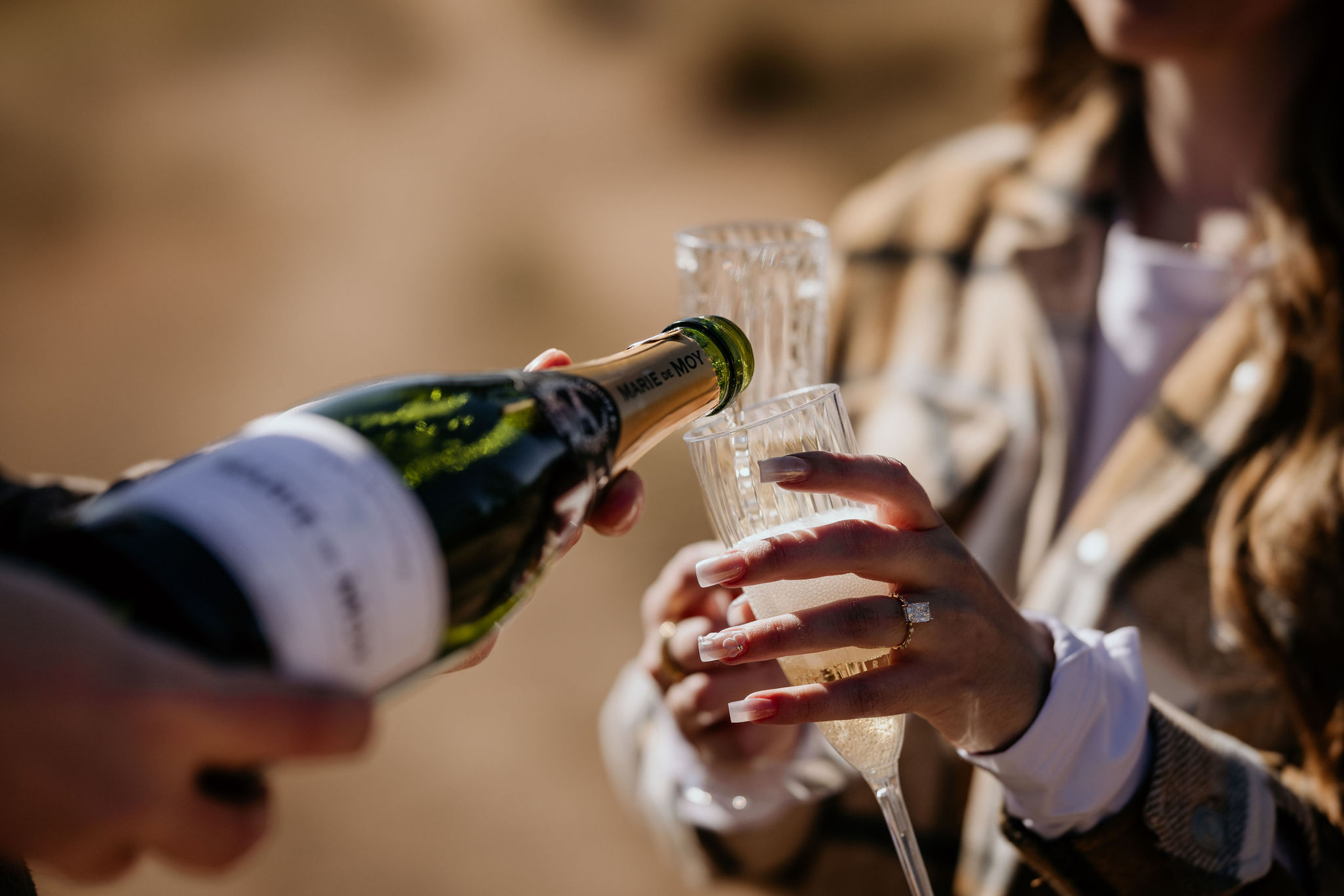 close up image of woman with engagement ring holding a glass and her fiancee pours champagne.