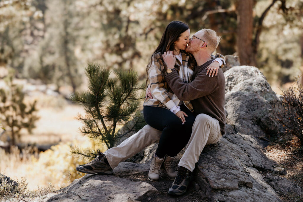 man and woman sit on rock and kiss during engagement photos.