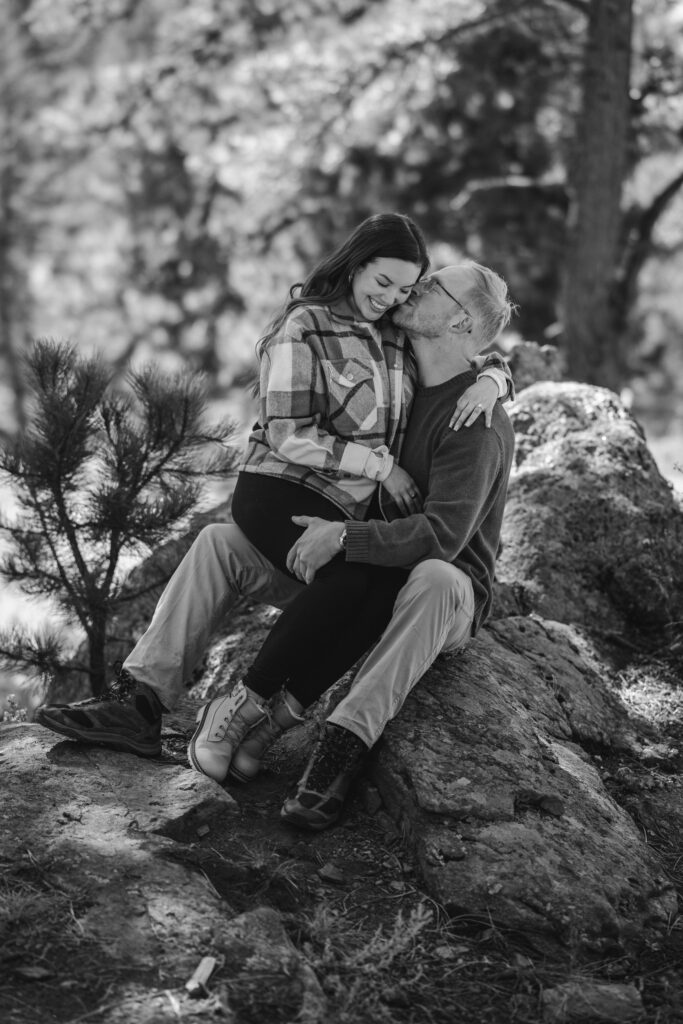 mand and woman sit on rock as he kisses her cheek during engagement photos.