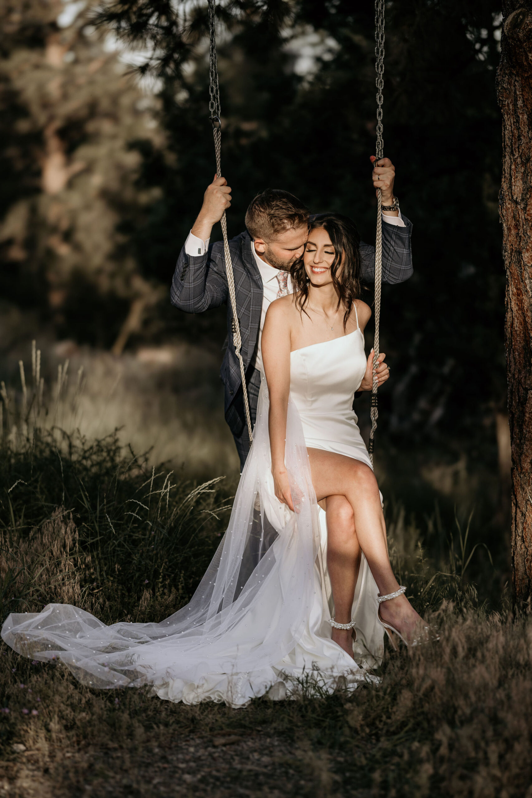 bride sits on tree swing while groom kisses her.