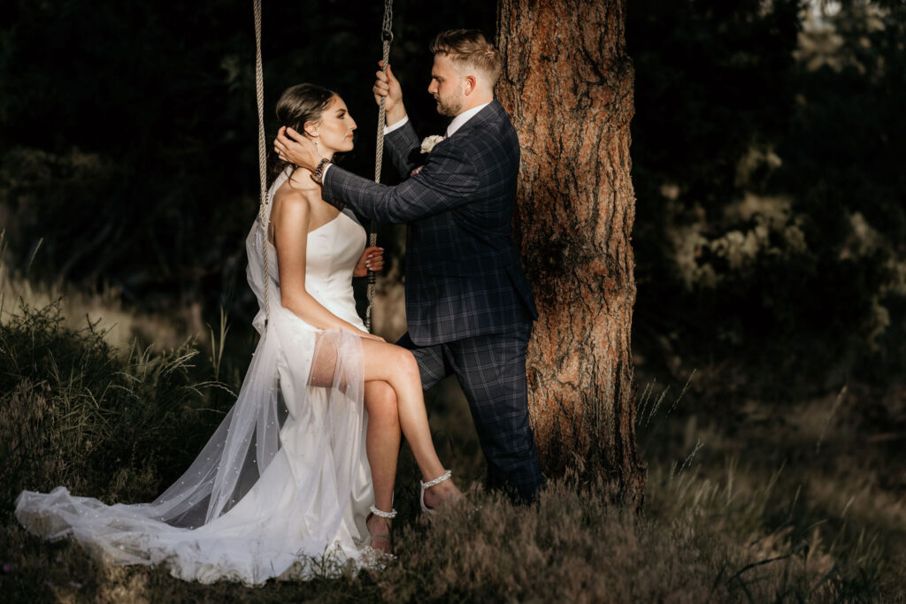 bride sits on tree swing while groom pushes hair out of her face during wedding photos.