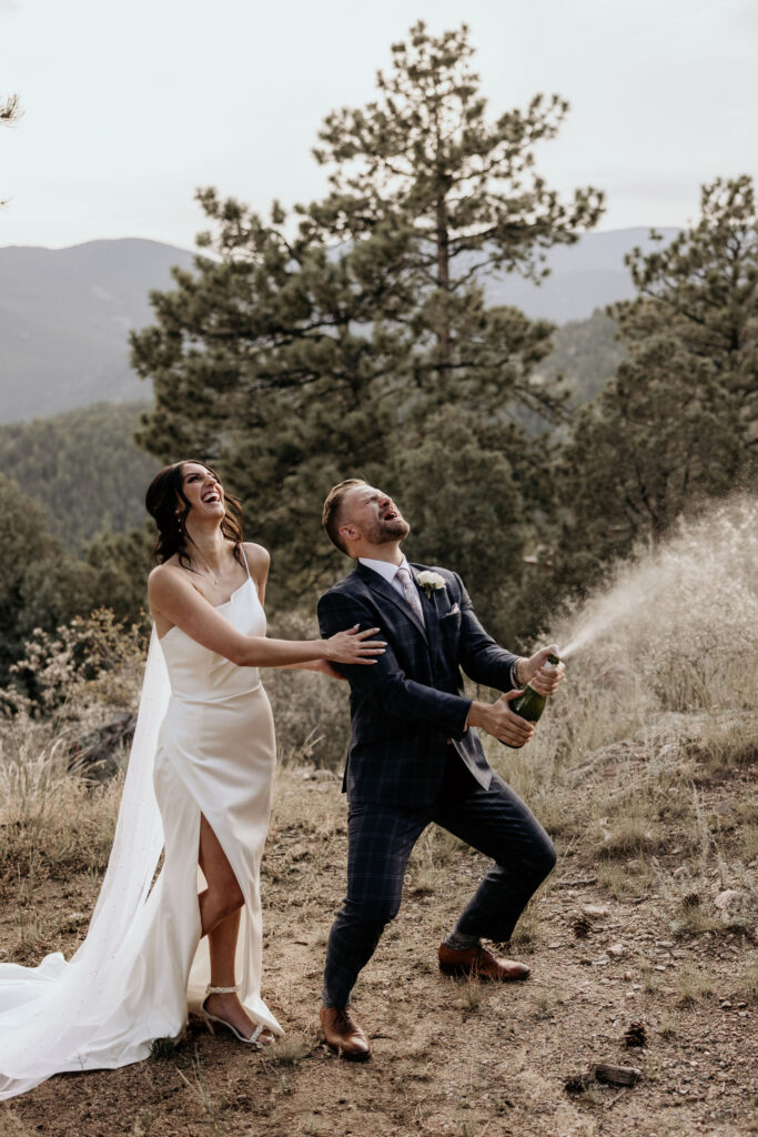 bride and groom laugh as they open champagne in the colorado mountains.