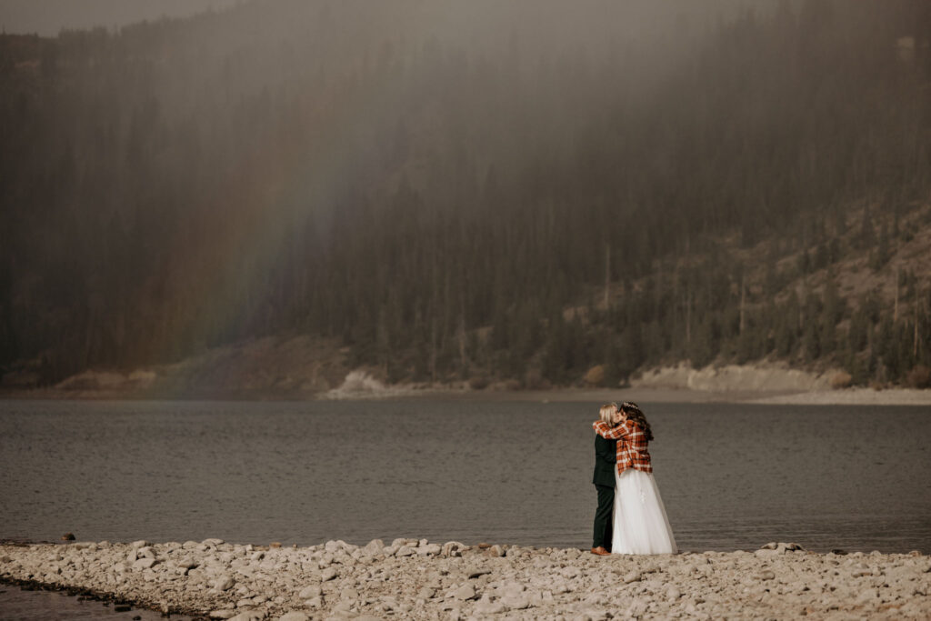 newlywed couple hugs with rainbow and mountains in the background during LGBTQ+ elopement.