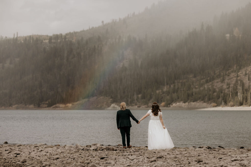 newlywed couple hold hands and look out at the mountains and a rainbow in breckenridge colorado.