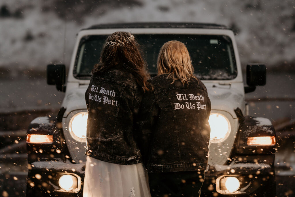 newlywed couple stand facing their jeep with matching jackets during snowy elopement photos.