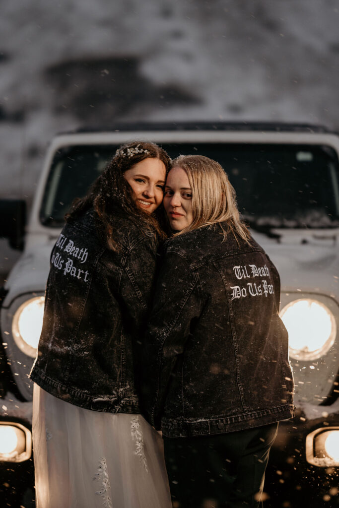 newlywed couple stands in front of jeep with matching jackets and smile during blue hour photos.