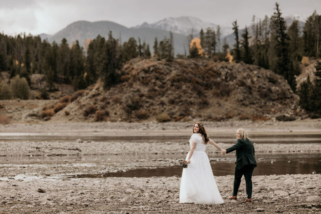 newlywed couple holds hands and walk on lake shore with colorado mountains in the background.
