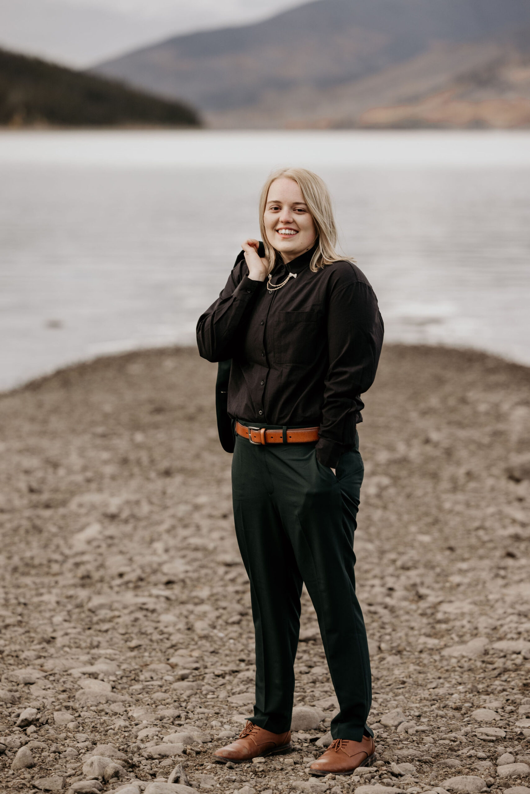 person in green and black suit stand in front of mountain lake during wedding portraits.