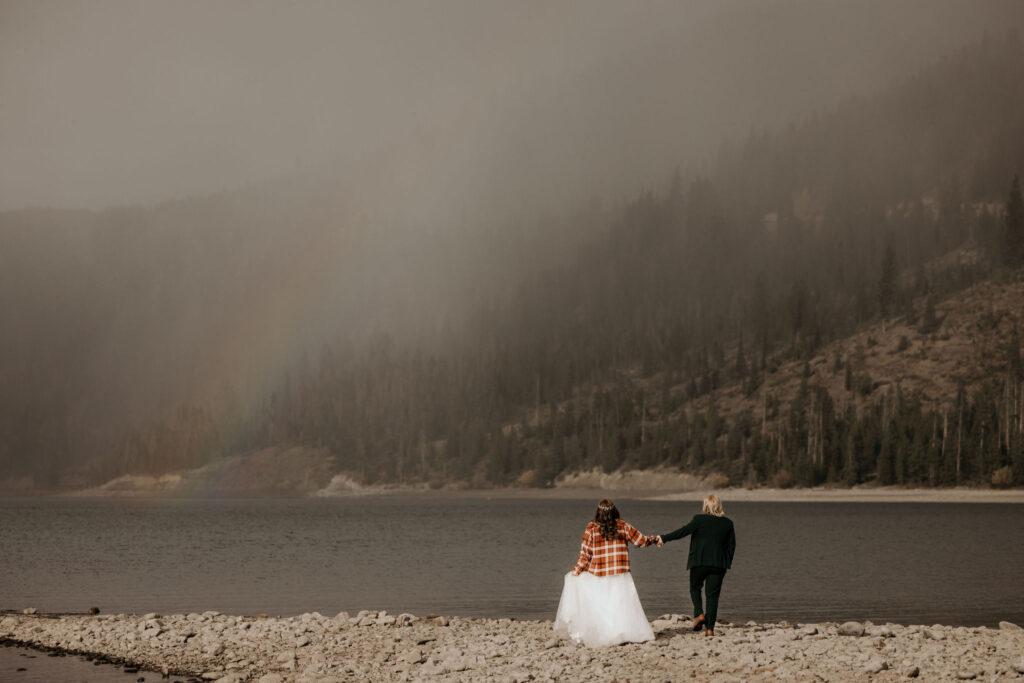 newlywed couple hold hands and walk along lake dillon during their LGBTQ+ wedding day.