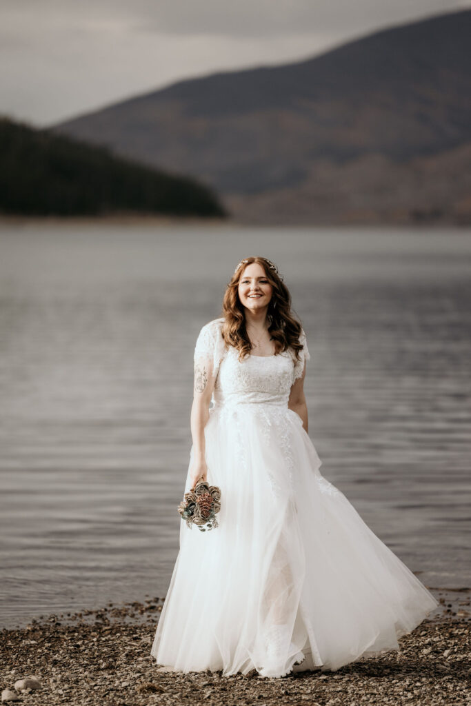 bride holds bouquet and stands in front of mountain lake during Breckenridge elopement.