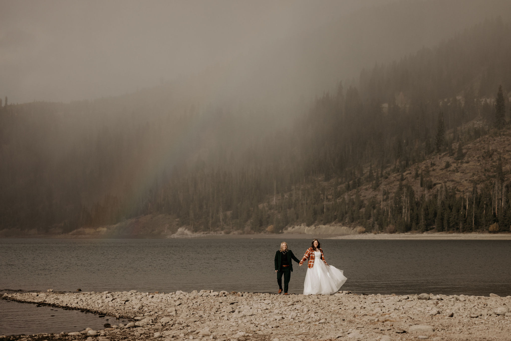 two brides holds hands with a rainbow in the background during their simple elopement in colorado.