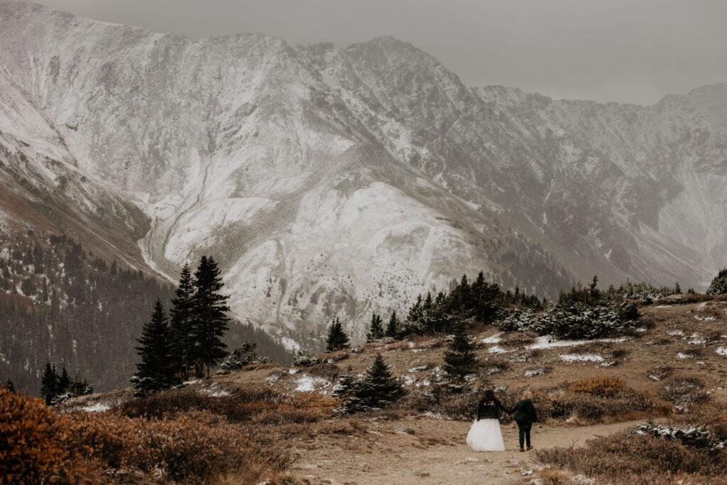 newlywed couple hold hands and stand on a trail looking at the snow covered mountains in breckenridge.