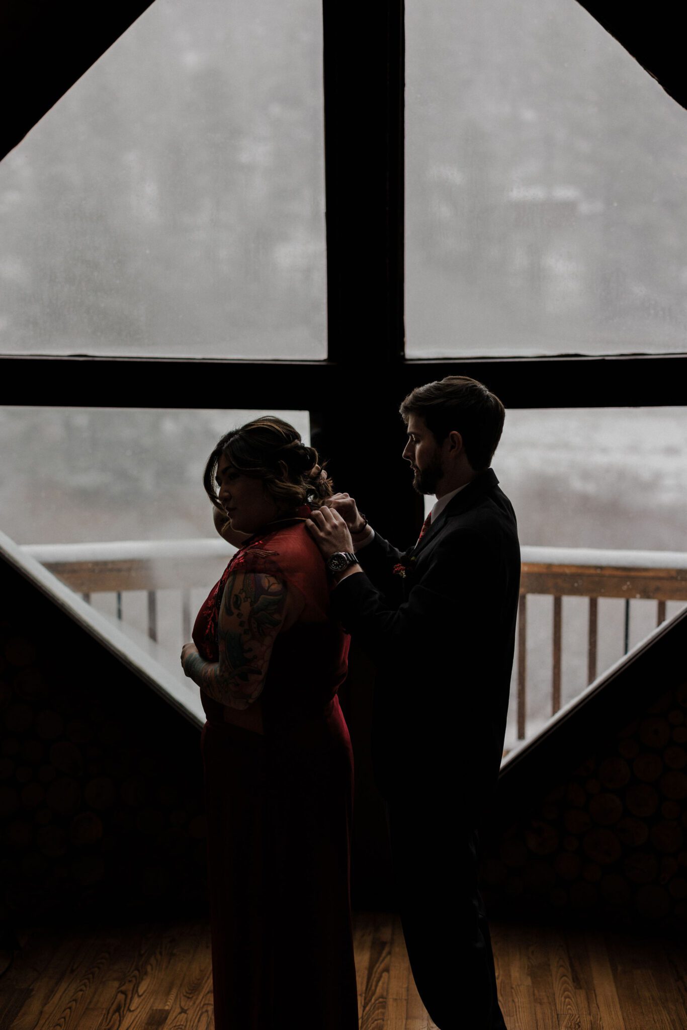 groom puts the bride's necklace on in front of a window looking out into the snow after grieving and planning a wedding