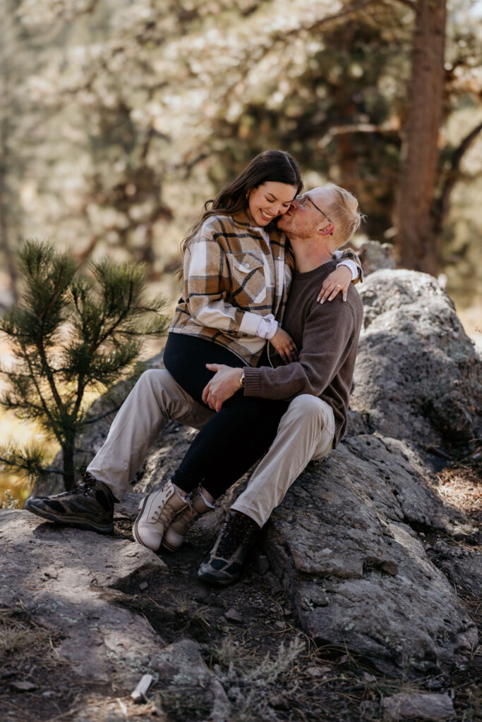 man and woman sit on rock while he kisses her cheek during engagement photos.